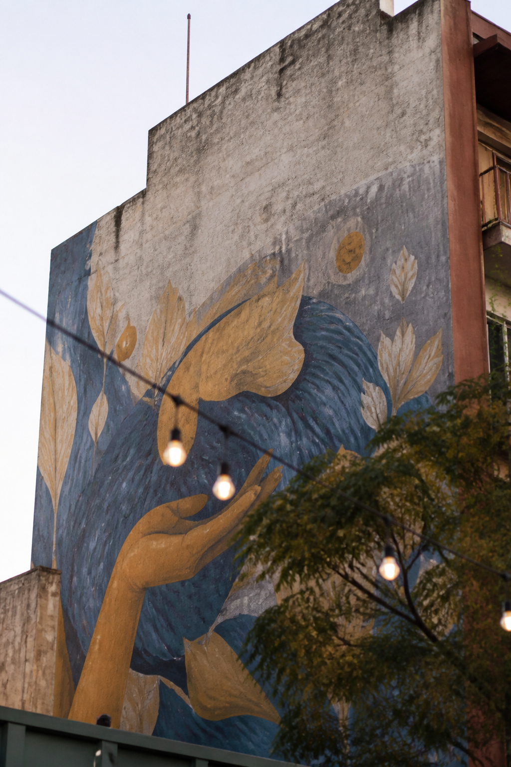Muro medianero de un edificio pintado con un mural de gran formato en ocres y azules, con cables de luz cruzando delante